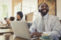 Lächelnder junger Mann mit Kopfhörern am Laptop und Kaffeetrinken am Cafétisch — Stockfoto