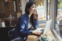 Giovane donna pensierosa che ascolta musica con cuffie al computer portatile e beve caffè nella finestra del caffè — Foto stock