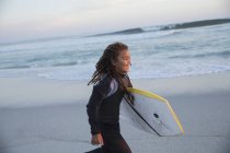 Smiling pre-adolescent girl in wet suit running with boogie board on summer beach — Stock Photo