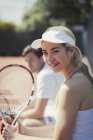 Портрет улыбающейся, уверенной в себе молодой теннисистки, держащей теннисную ракетку на солнечном теннисном корте — стоковое фото
