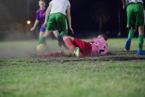Young female soccer player falling, kicking the ball playing soccer on field at night — Stock Photo