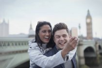 Веселая, любовная пара туристов, делающих селфи с телефоном перед Вестминстерским мостом, Лондон, Великобритания — стоковое фото