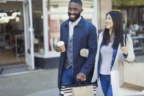 Sonriente pareja joven caminando brazo en brazo a lo largo de escaparate con café y bolsas de compras - foto de stock