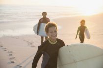 Retrato sorrindo menino pré-adolescente em terno molhado carregando prancha de surf na praia do pôr do sol verão com a família — Fotografia de Stock