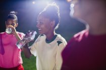Smiling young female soccer players resting, drinking from water bottles on field at night — Stock Photo
