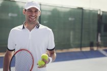 Уверенный в себе портрет, улыбающийся молодой теннисист, держащий теннисную ракетку и теннисные мячи на солнечном теннисном корте — стоковое фото