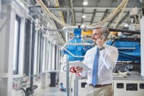 Supervisore maschile con tablet digitale che parla sul telefono cellulare accanto a macchinari per stampaggio industriale in plastica in fabbrica — Foto stock