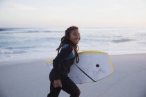 Portrait happy pre-adolescent girl running with boogie board on summer beach at dusk — Stock Photo