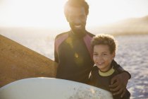 Портрет улыбающийся, уверенный в себе отец и сын в мокрых костюмах с досками для серфинга на солнечном летнем пляже — стоковое фото