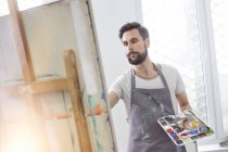 Male artist with palette painting at easel in art studio — Stock Photo