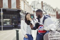 Молодая пара смеется, используя мобильный телефон на мотороллере на солнечной городской улице — стоковое фото