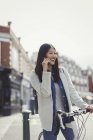 Lächelnde junge Frau, die mit dem Fahrrad pendelt, auf der sonnigen Stadtstraße mit dem Handy telefoniert — Stockfoto