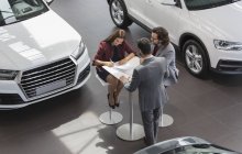 Car salesman watching couple customers signing financial contract paperwork in car dealership showroom — Stock Photo