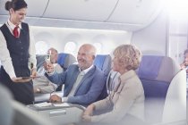 Auxiliar de vuelo sirviendo champán a pareja madura en primera clase en avión - foto de stock