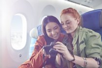 Smiling young women friends looking at photos on digital camera on airplane — Stock Photo