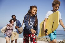 Sonriente familia llevando boogie board, caminando por soleado sendero de playa de verano - foto de stock
