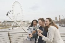 Smiling friend tourists celebrating, toasting champagne and taking selfie with selfie stick near Millennium Wheel, London, UK — Stock Photo