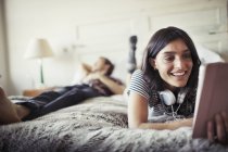 Улыбающаяся молодая женщина в наушниках с помощью цифрового планшета на кровати — стоковое фото
