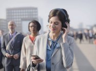 Geschäftsfrau lächelt, hört Musik mit Kopfhörer und Smartphone auf städtischer Fußgängerbrücke — Stockfoto