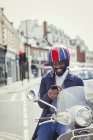 Улыбающийся молодой бизнесмен в шлеме на мотороллере, переписывающийся с сотовым на городской улице — стоковое фото