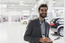 Retrato sonriente, vendedor de coches con confianza portapapeles en concesionario de automóviles taller de reparación de automóviles - foto de stock