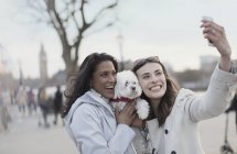 Pareja lésbica juguetona y sonriente con perro blanco tomando selfie con teléfono de cámara en el parque urbano, Londres, Reino Unido - foto de stock