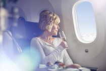 Mujer madura bebiendo champán, mirando por la ventana en primera clase en el avión - foto de stock
