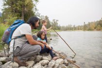 Отец и дочери рыбачат палками на берегу озера. — стоковое фото