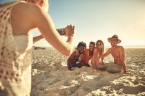 Молода жінка з телефоном фотографує друзів на сонячному літньому пляжі — стокове фото