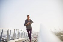 Улыбающаяся бегунья проверяет умные часы фитнес-трекер на солнечном городском мосту — стоковое фото