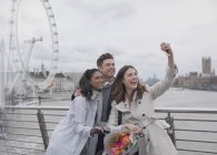 Smiling, happy friends taking selfie with selfie stick on bridge near Millennium Wheel, London, UK — Stock Photo