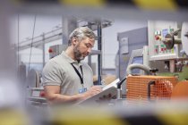 Superviseur masculin avec presse-papiers au panneau de commande des machines dans l'usine de fibre optique — Photo de stock