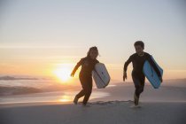 Bruder und Schwester im Neoprenanzug laufen mit Boogie Boards am Strand des sommerlichen Sonnenuntergangs — Stockfoto
