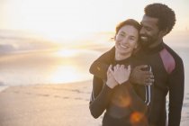 Smiling, affectionate multi-ethnic couple in wet suits on sunny summer sunset beach — Stock Photo
