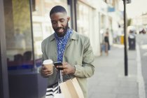 Young man with coffee and shopping bags texting with cell phone on urban sidewalk — Stock Photo