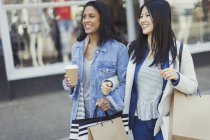Mujeres sonrientes amigas caminando brazo en brazo a lo largo del escaparate con café y bolsas de compras - foto de stock