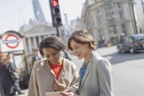 Femmes d'affaires souriantes avec tablette numérique parlant sur la rue urbaine ensoleillée — Photo de stock