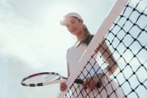 Retrato sorrindo, jogador de tênis feminino confiante segurando raquete de tênis na rede abaixo do céu ensolarado — Fotografia de Stock