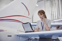 Женщина-инженер самолета работает за ноутбуком и разговаривает по мобильному телефону в ангаре — стоковое фото
