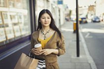 Young woman walking along storefront with coffee cup and shopping bags — Stock Photo