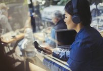 Молодая женщина слушает музыку в наушниках, пишет смс с мобильного телефона у окна кафе — стоковое фото