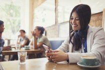 Smiling young woman with headphones texting with cell phone and drinking coffee at cafe table — Stock Photo