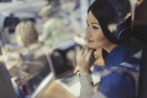 Mujer joven pensativa en el ordenador portátil escuchando música con auriculares en la ventana de la cafetería - foto de stock
