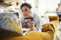 Jeune femme avec casque et carte de crédit achats en ligne à l'ordinateur portable sur le canapé du salon — Photo de stock