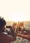 Young man with camera phone photographing friends enjoying picnic on sunny summer beach — Stock Photo