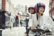 Smiling young women friends wearing helmets and riding motor scooter on urban street — Stock Photo