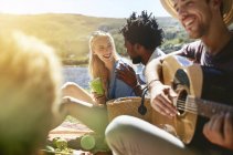 Jóvenes amigos tocando la guitarra y disfrutando de un soleado picnic de verano - foto de stock