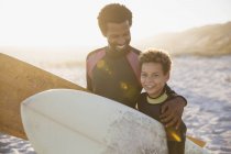 Retrato sonriente padre e hijo surfistas llevando tablas de surf en la soleada playa de verano - foto de stock