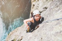 Escalador femenino enfocado y determinado escalando rocas por encima del océano soleado - foto de stock