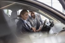 Vendedor de coches explicando coche nuevo a cliente femenino en el asiento del conductor en la sala de exposición de concesionarios de automóviles - foto de stock